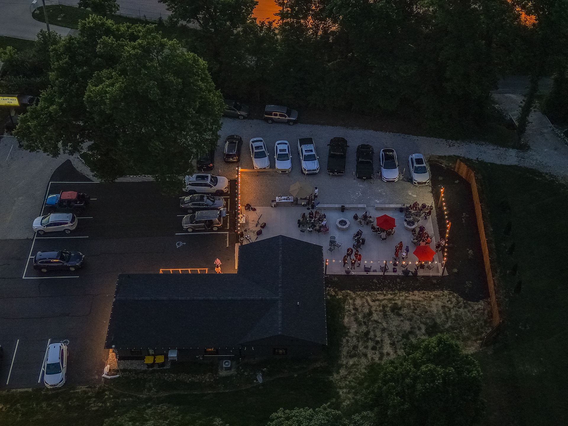 Evening aerial view of The Sip Craft Beer Pub showing the building, parking, illuminated patio, and guests